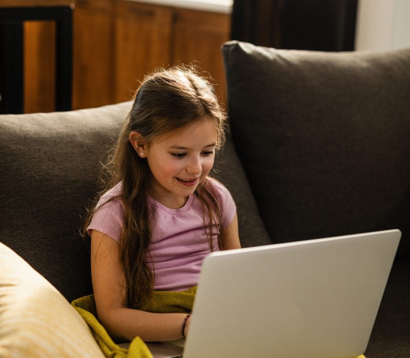 Smiling little girl sitting on the sofa and using laptop in the living room.